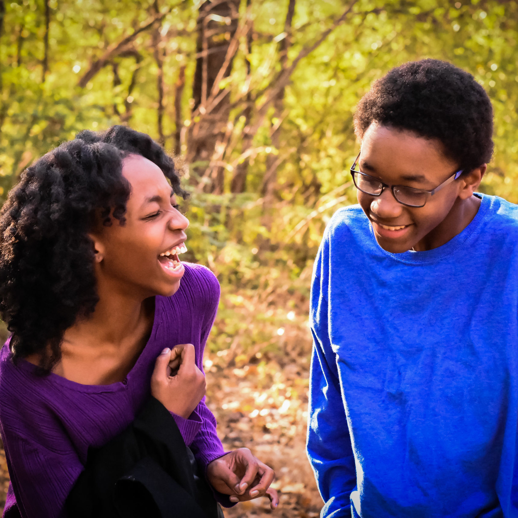 Two siblings walk in the forest and laugh hard. Sister on the left faces brother on the right with an open mouth grin.