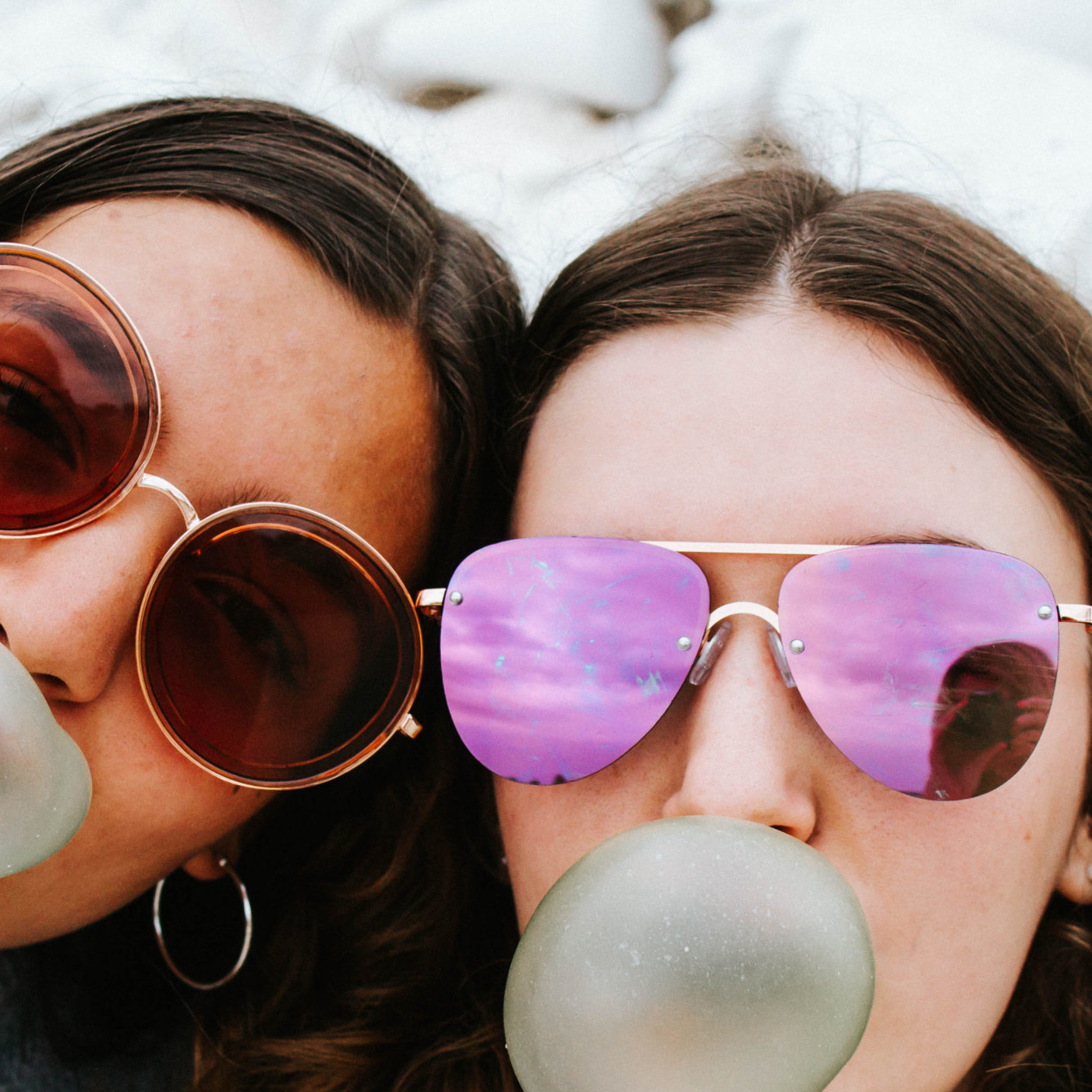 Two teen girls wearing sunglasses blow bubble gum bubbles.