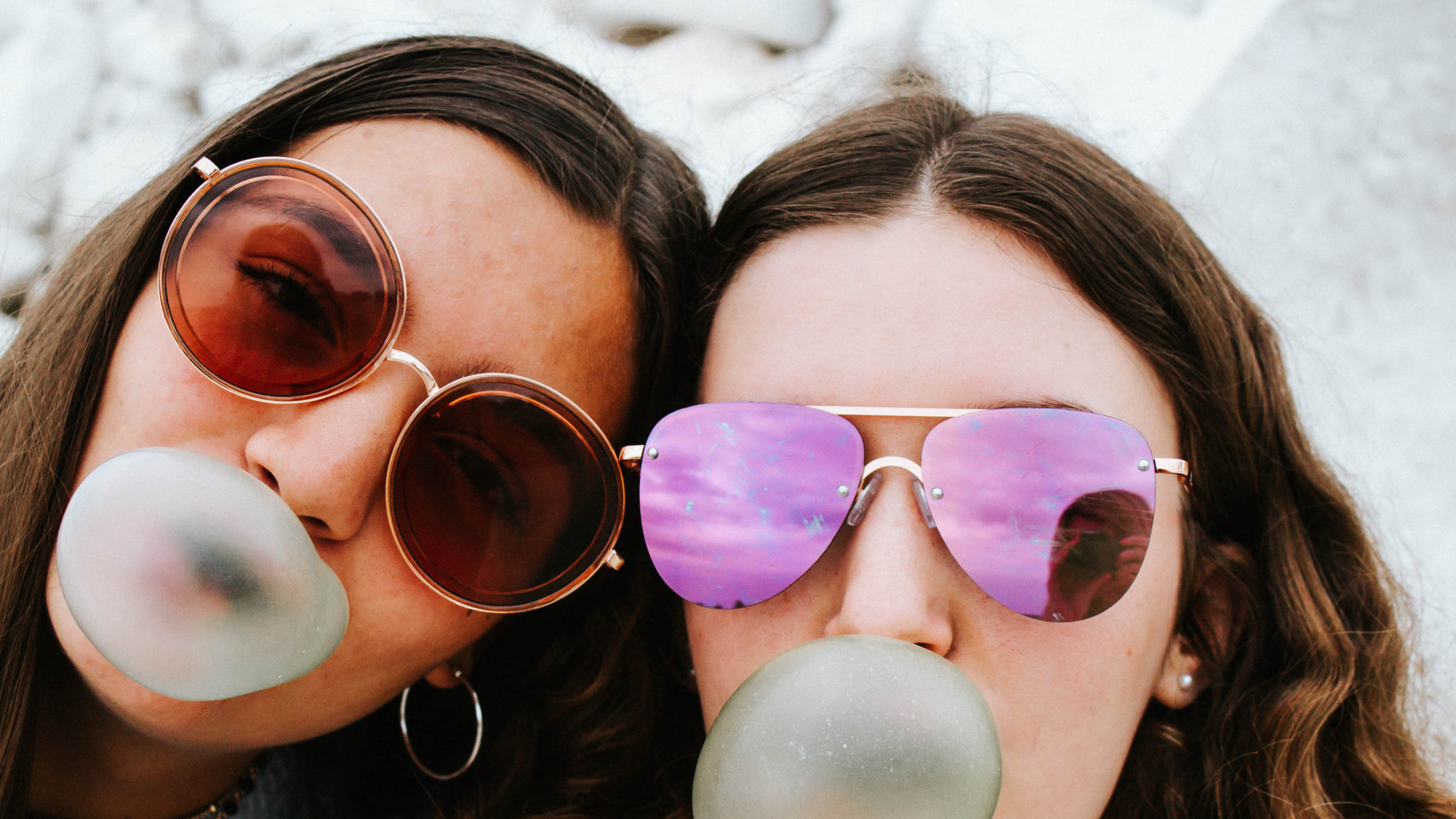 Two teen girls wearing sunglasses blow bubble gum bubbles.