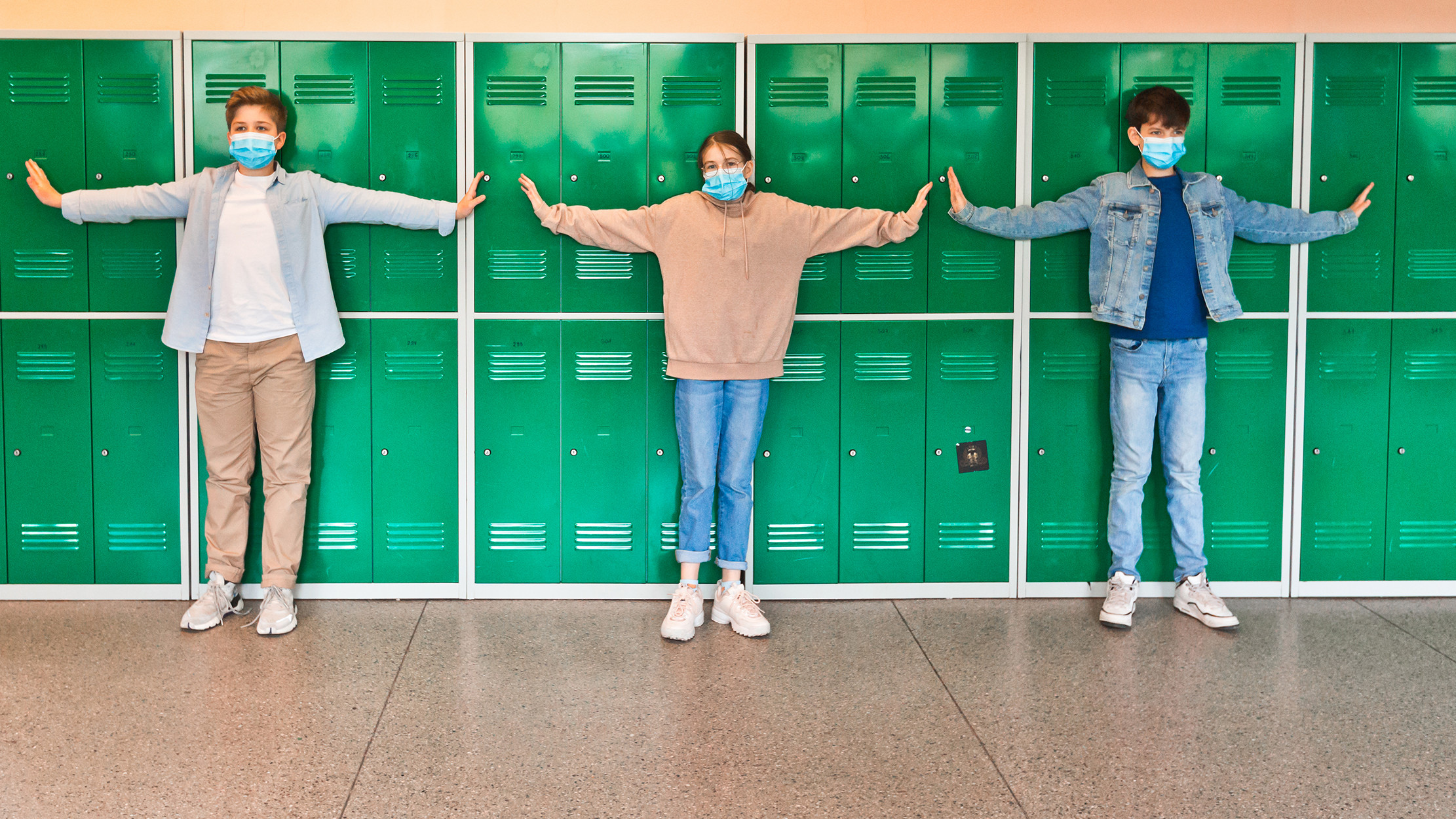 Teenage boys and girl wearing N95 face masks standing in front of school lockers and outstretching arms in the distance. High school students at school.