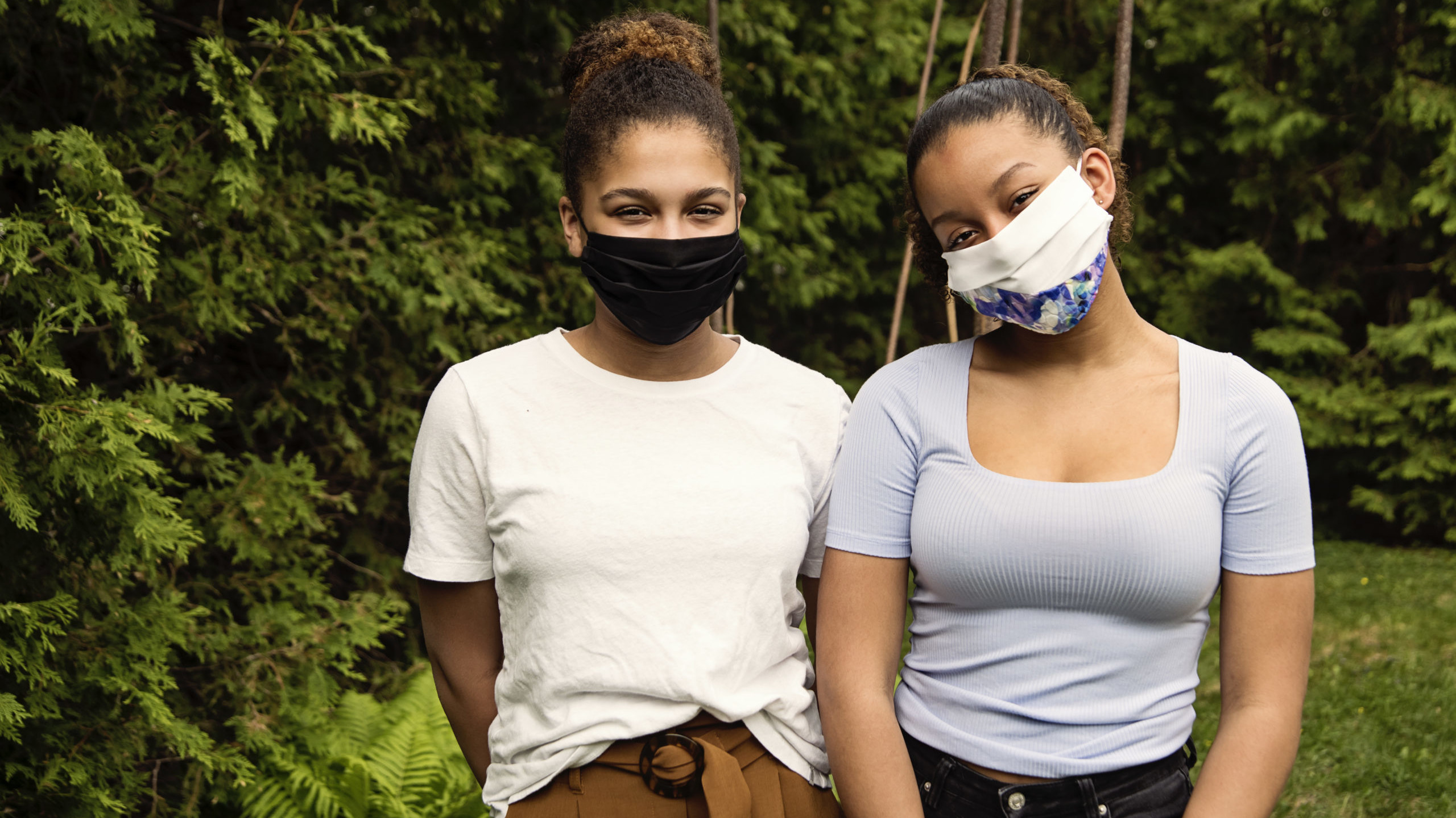 Two teenage sisters smiling behind their stylish protective black mask. They are mixed-race and looking at the camera. Nature in background. Horizontal outdoors waist up shot with copy space.
