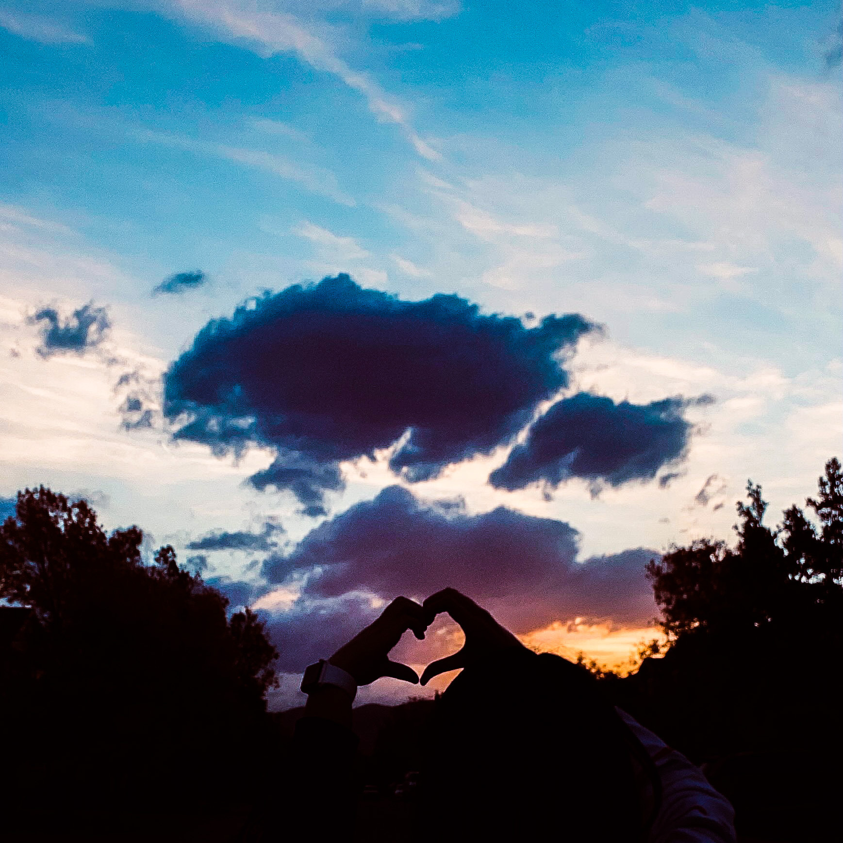 A colorful sunset covers the sky with two half-heart hands connecting to make a heart shape in front of the sky.