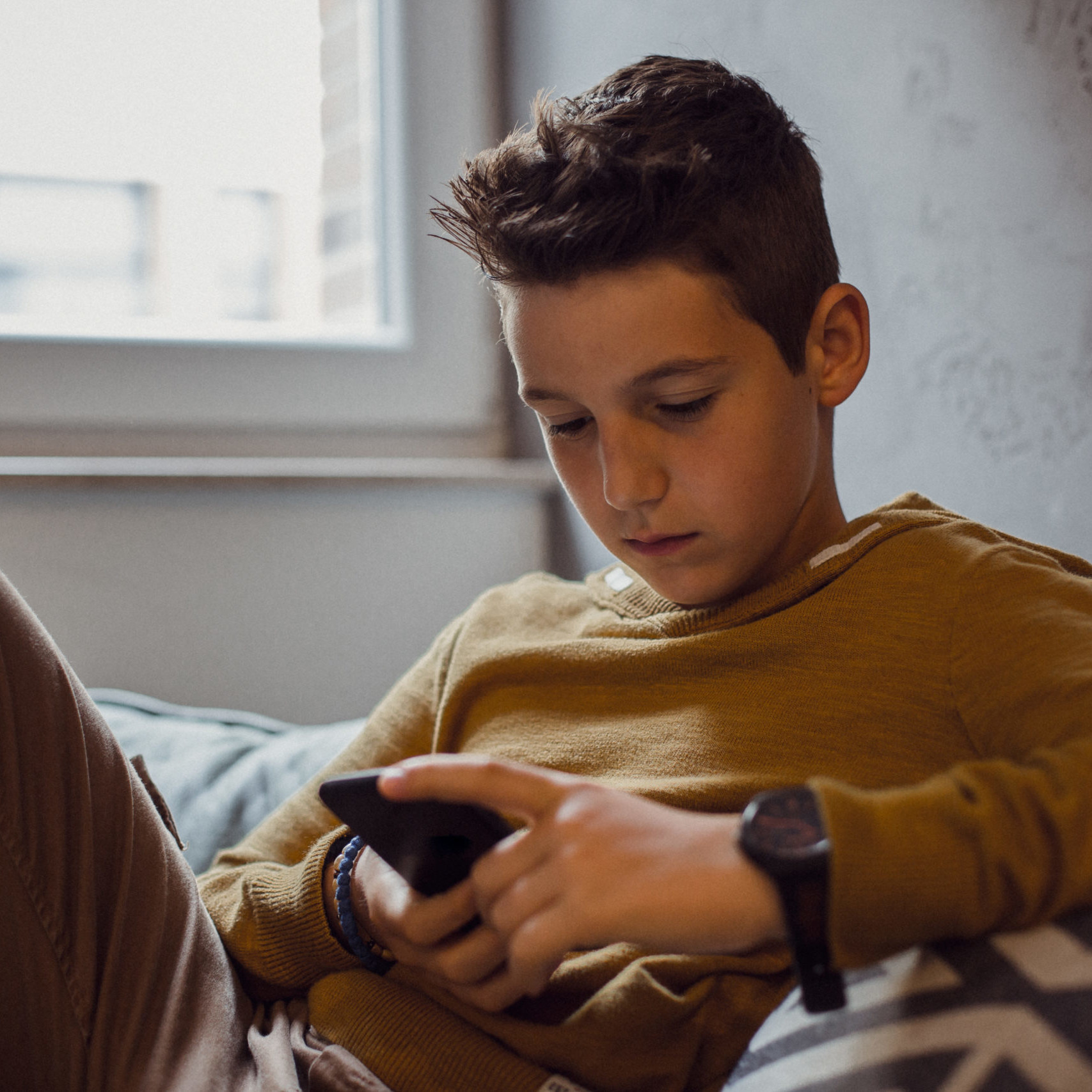 Young boy sitting on his bed and looking at his phone.
