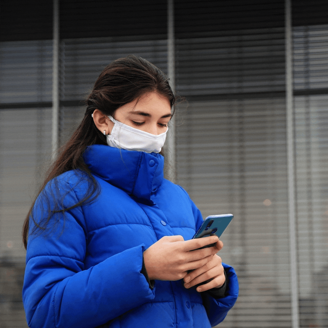 Teen girl wearing a blue winter jacket and a face mask walks and scrolls on her phone outdoors.